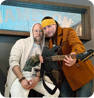 Two men posing together, one holding an electric guitar
